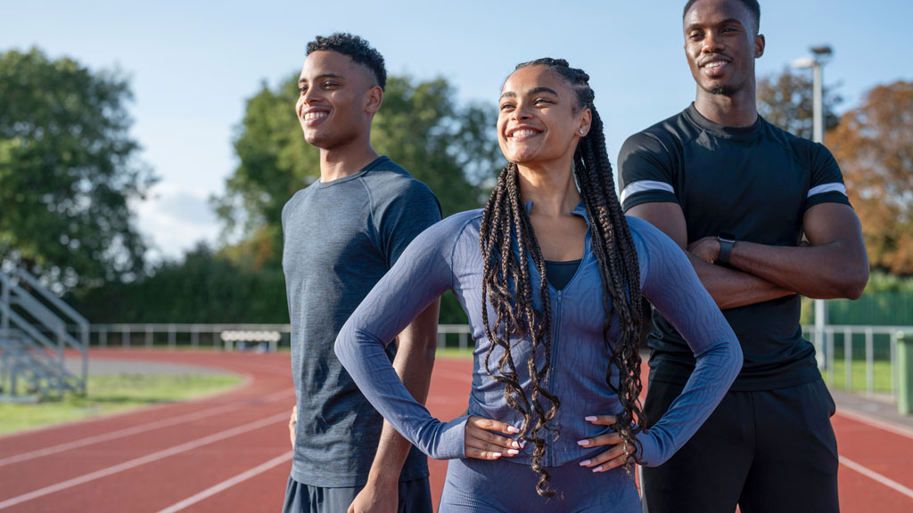 Groupe d'athlètes universitaires divers s'échauffant sur terrain de sport avec installations campus visibles